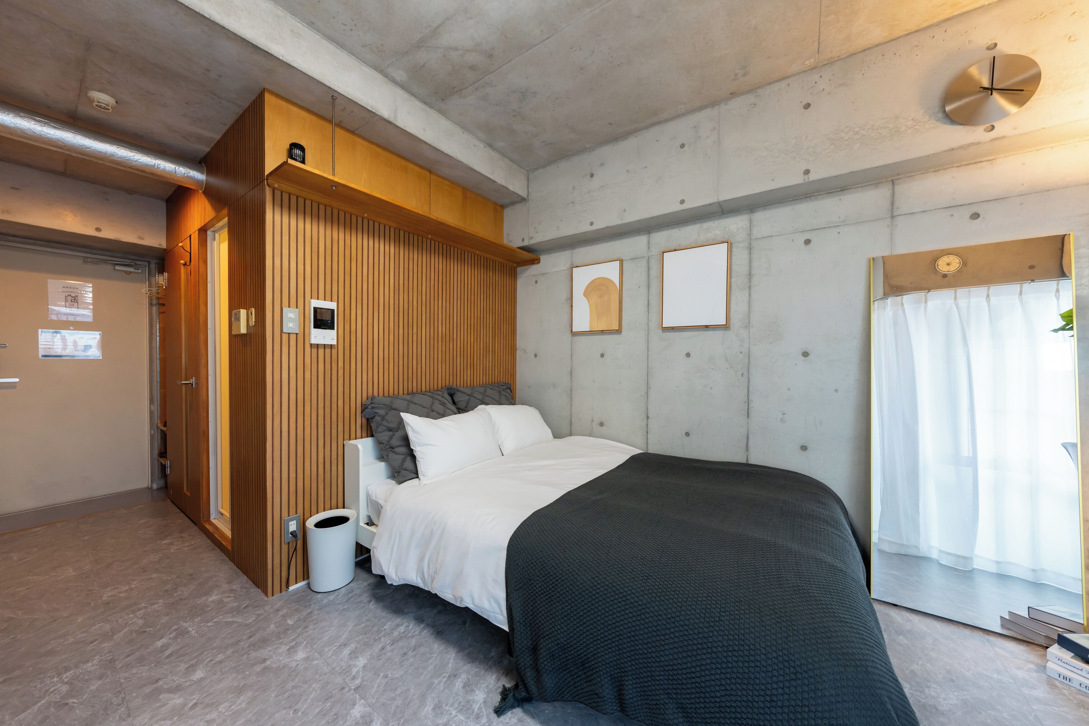A bedroom with a bed, wooden paneling, a white wall clock, and a window with sheer curtains.