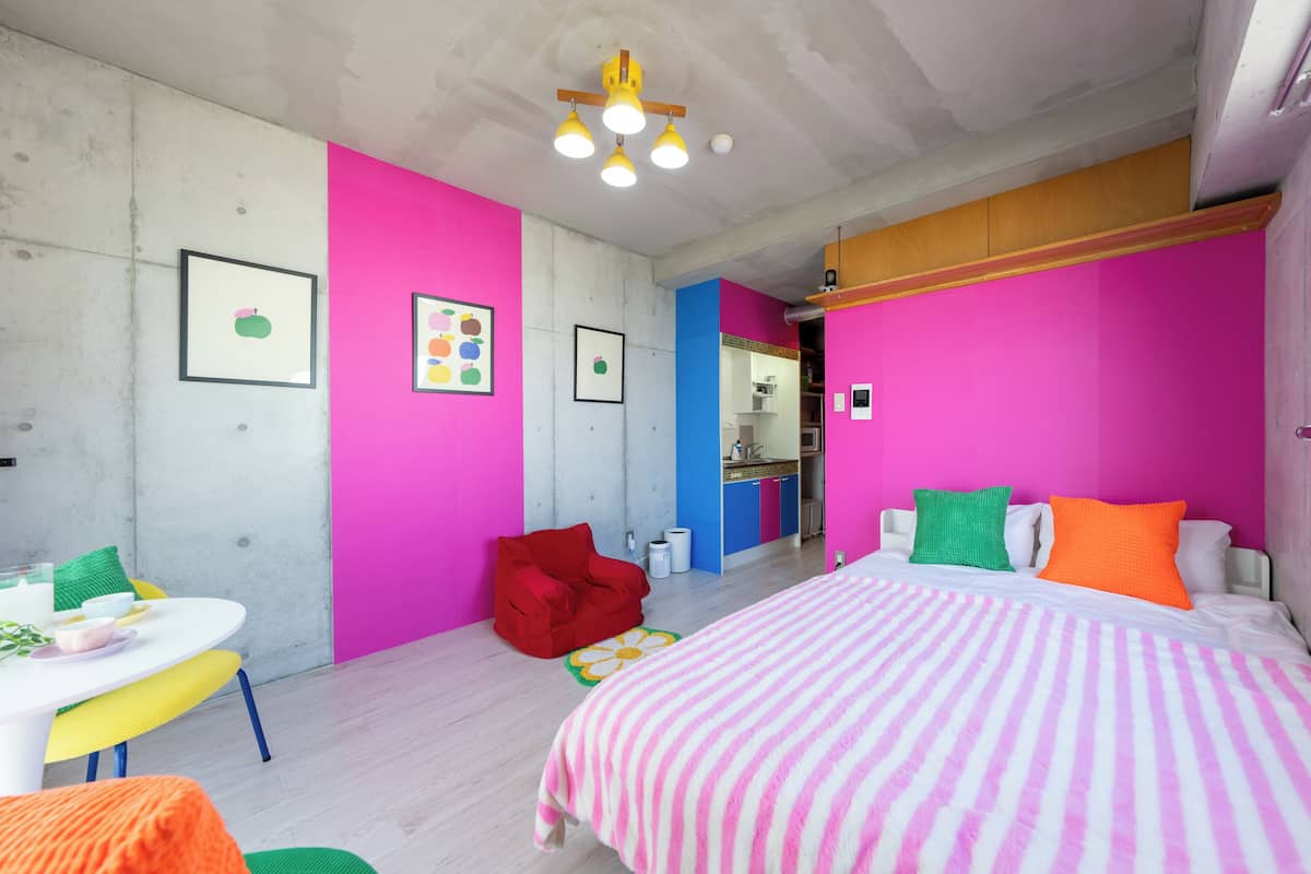 A modern bedroom with a bed, a red chair, a small table with a plant, and a kitchenette in the background.