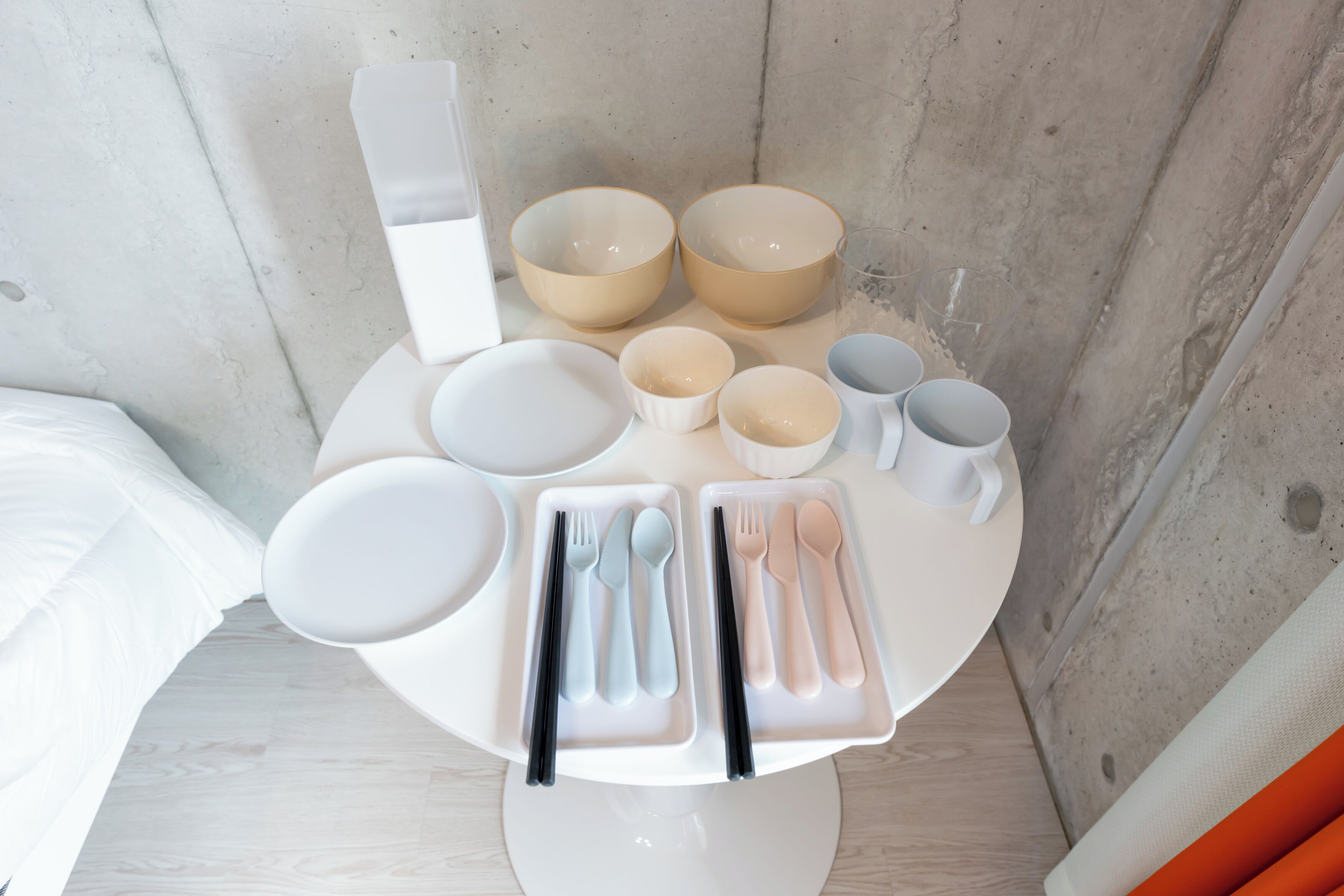 Un juego de comedor con cuencos, platos y cubiertos sobre una mesa blanca frente a una pared de concreto.