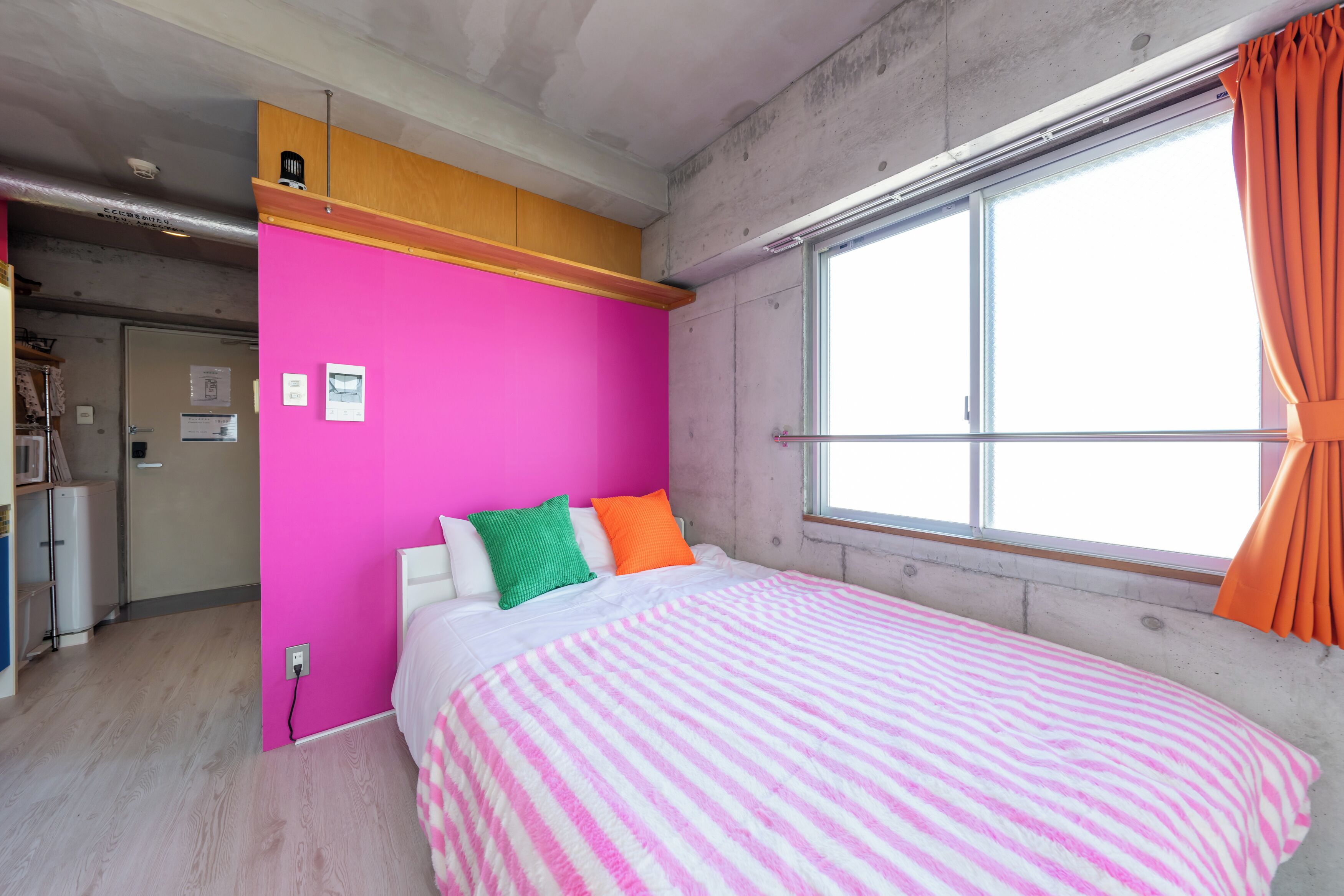 A bedroom with a pink wall, a bed with a striped blanket, and a window with orange curtains.