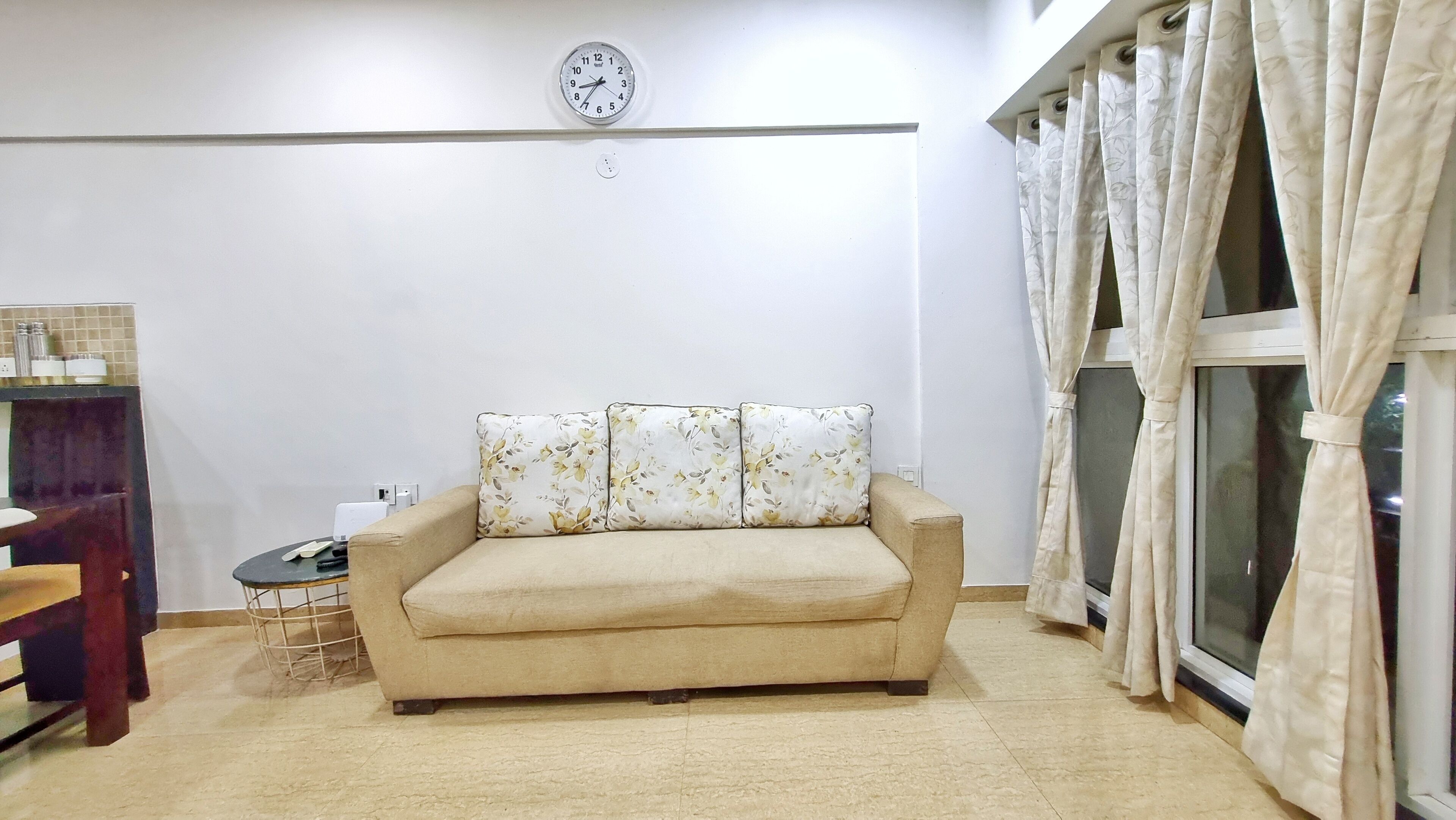 A beige sofa with floral cushions, a clock on the wall, and a window with curtains.