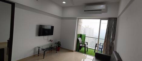 A modern living room with a flat-screen TV, a glass coffee table, a sofa, and a balcony with a view of high-rise buildings.