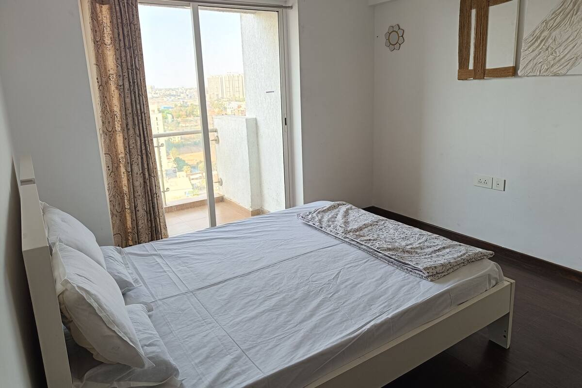 A bedroom with a bed, a window with curtains, a view of buildings, and a wall with a mirror and a light switch.