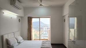 Ein Schlafzimmer mit Bett, Deckenventilator, Klimaanlage und einem Fenster mit Blick auf Gebäude und Berge.