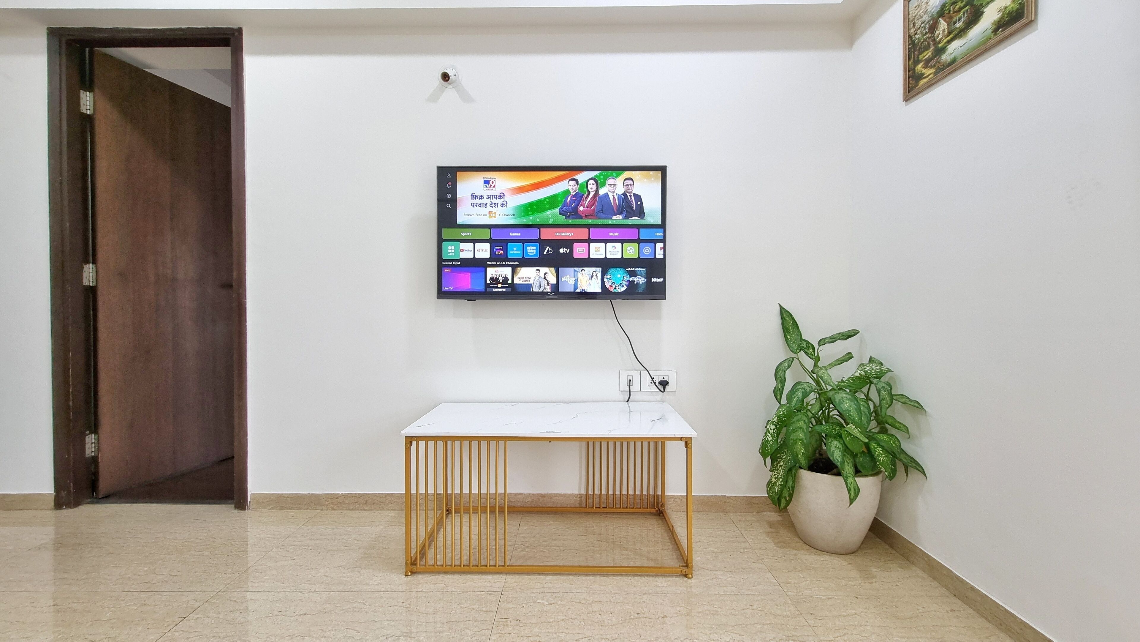 A room with a flat-screen TV mounted on the wall displaying various channels, a small wooden table in the center, and a potted plant on the right.