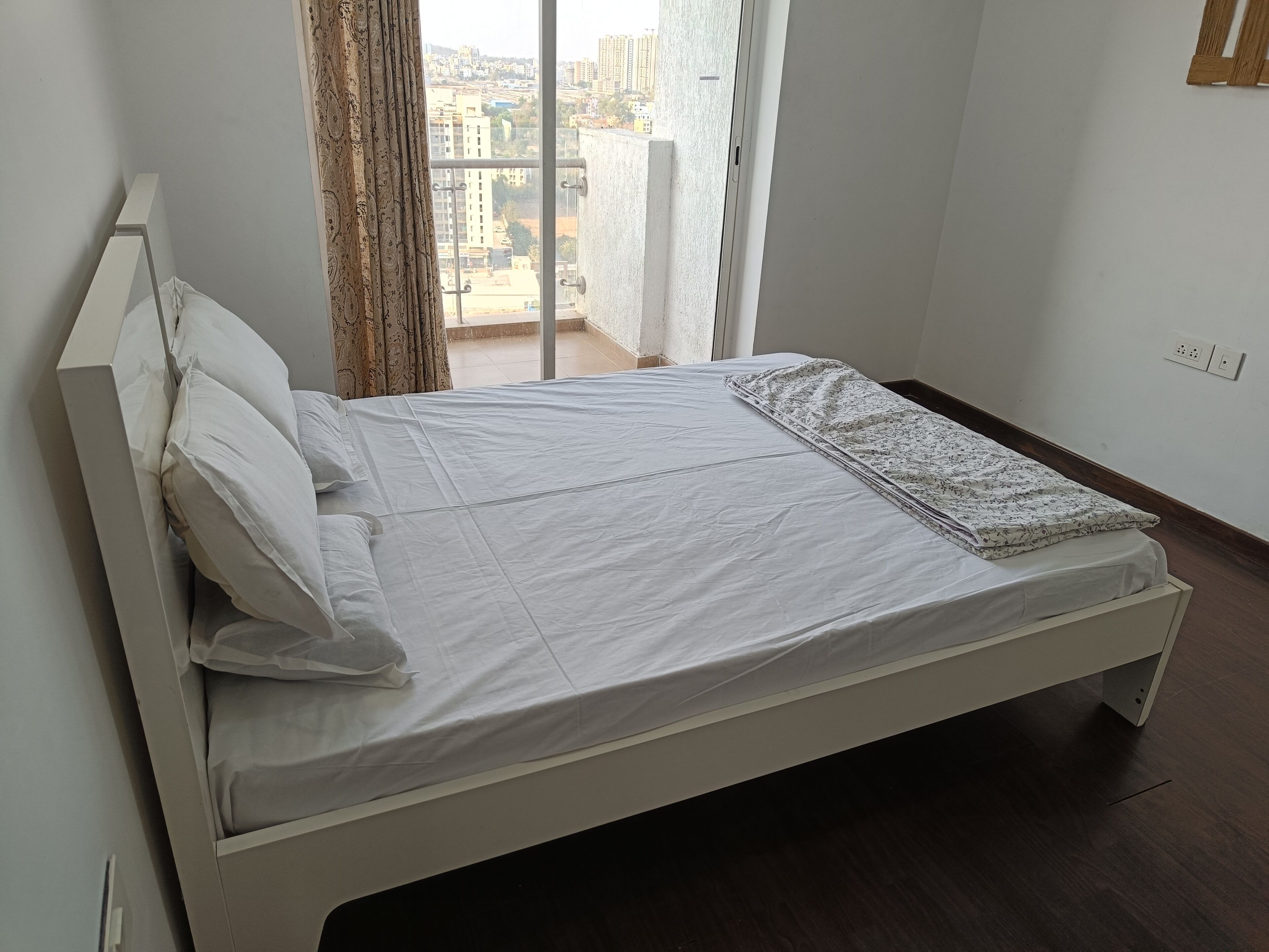 A single-bed room with a white bedspread, a patterned blanket, and a view of a cityscape through an open window.
