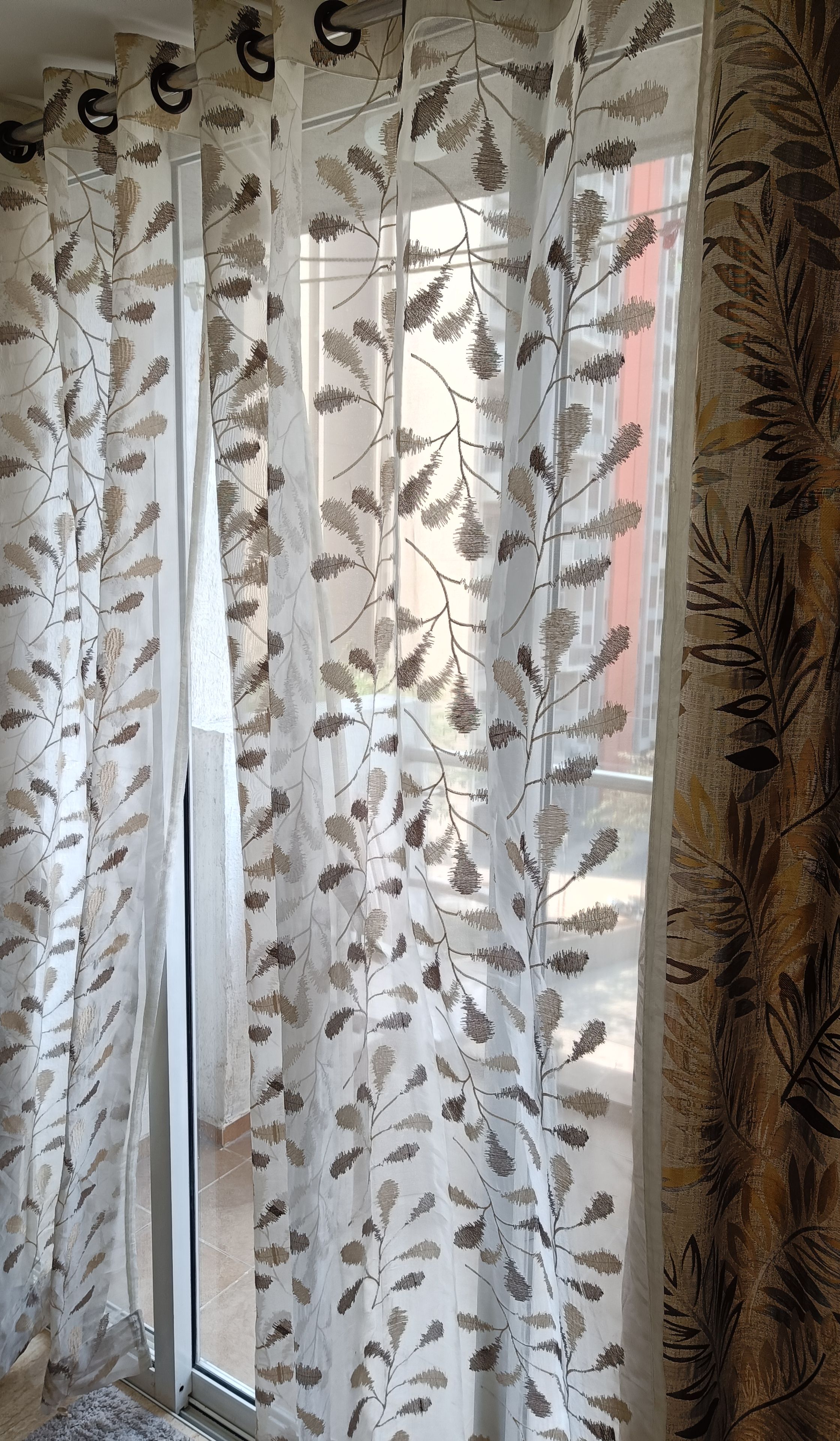 A room with sheer curtains featuring a leaf pattern, a sliding glass door, and a patterned fabric on the right side.