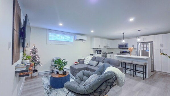 A modern living room with a grey sofa, a coffee table, and a small potted plant. There is a kitchen area with white cabinets and a stainless steel refrigerator. - Elegant Modern Loft in Prestigious Fairways (Riverview)