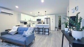 A modern kitchen with white cabinets, a central island, and stainless steel appliances. There is a grey sofa with cushions in the living area. - Elegant Modern Loft in Prestigious Fairways (Riverview)