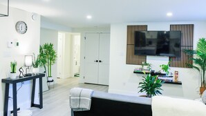 A modern living room with a flat-screen TV mounted on the wall, a black console table with a lamp, and several potted plants. - Elegant Modern Loft in Prestigious Fairways (Riverview)