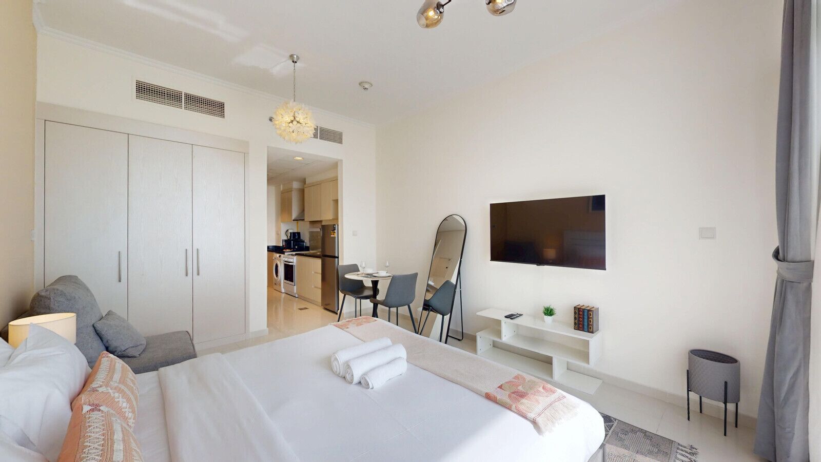 A modern hotel room with a large bed, a desk, a chair, a TV, and a kitchenette in the background.