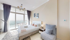 A hotel room with a large bed, a gray armchair, a nightstand with a lamp, and a view of the sea through large windows.