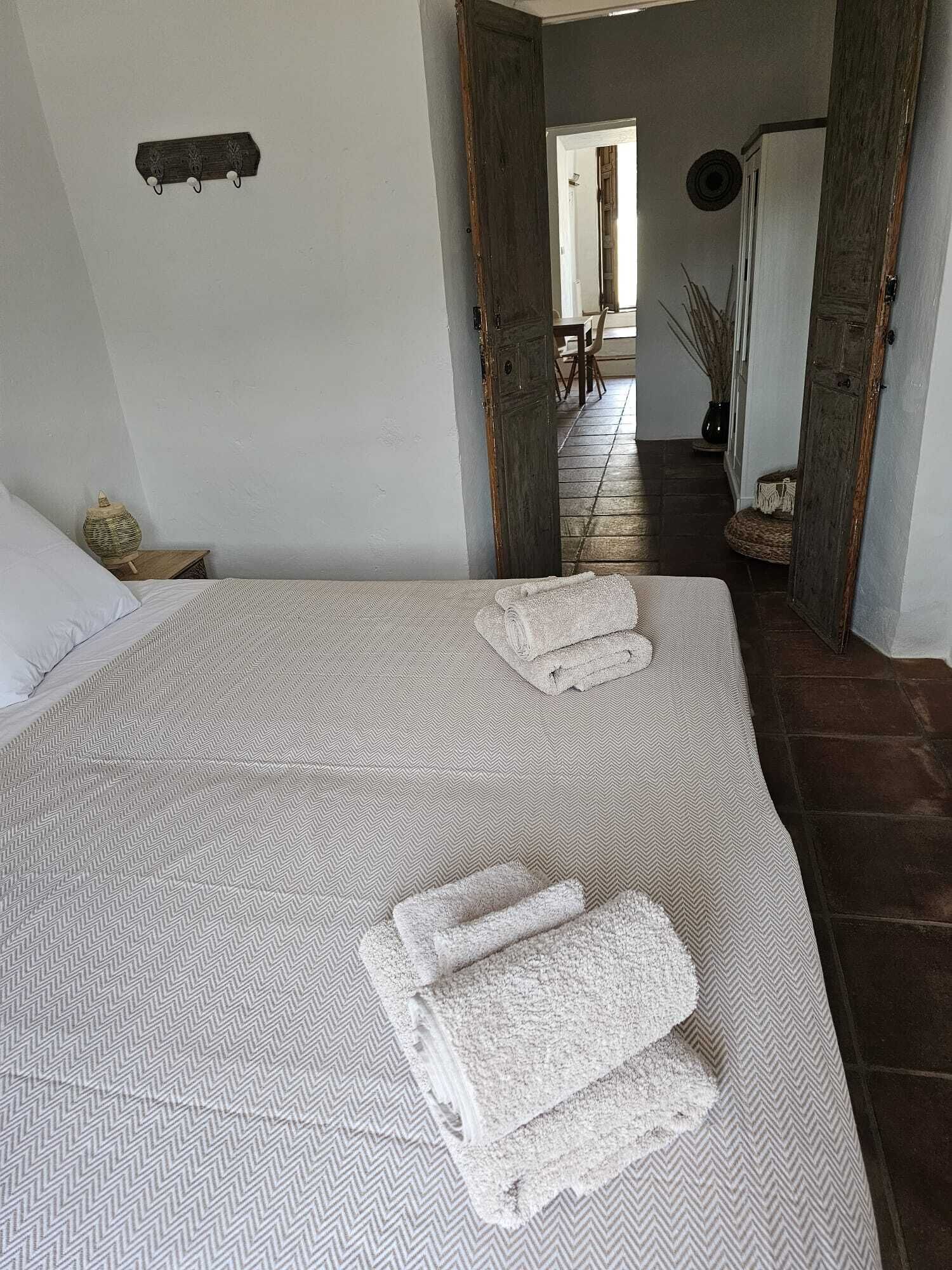 A bedroom with a large bed, two rolled towels, a small bedside table, and a wall-mounted shelf.