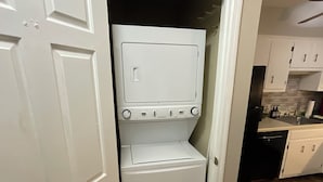 Una safareig petit amb rentadora i assecadora, una porta blanca, i una zona de cuina amb armaris i un taulell.