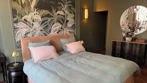 Un dormitorio con cama, mesita de noche, lámpara, espejo y una pared con un mural de plantas.
