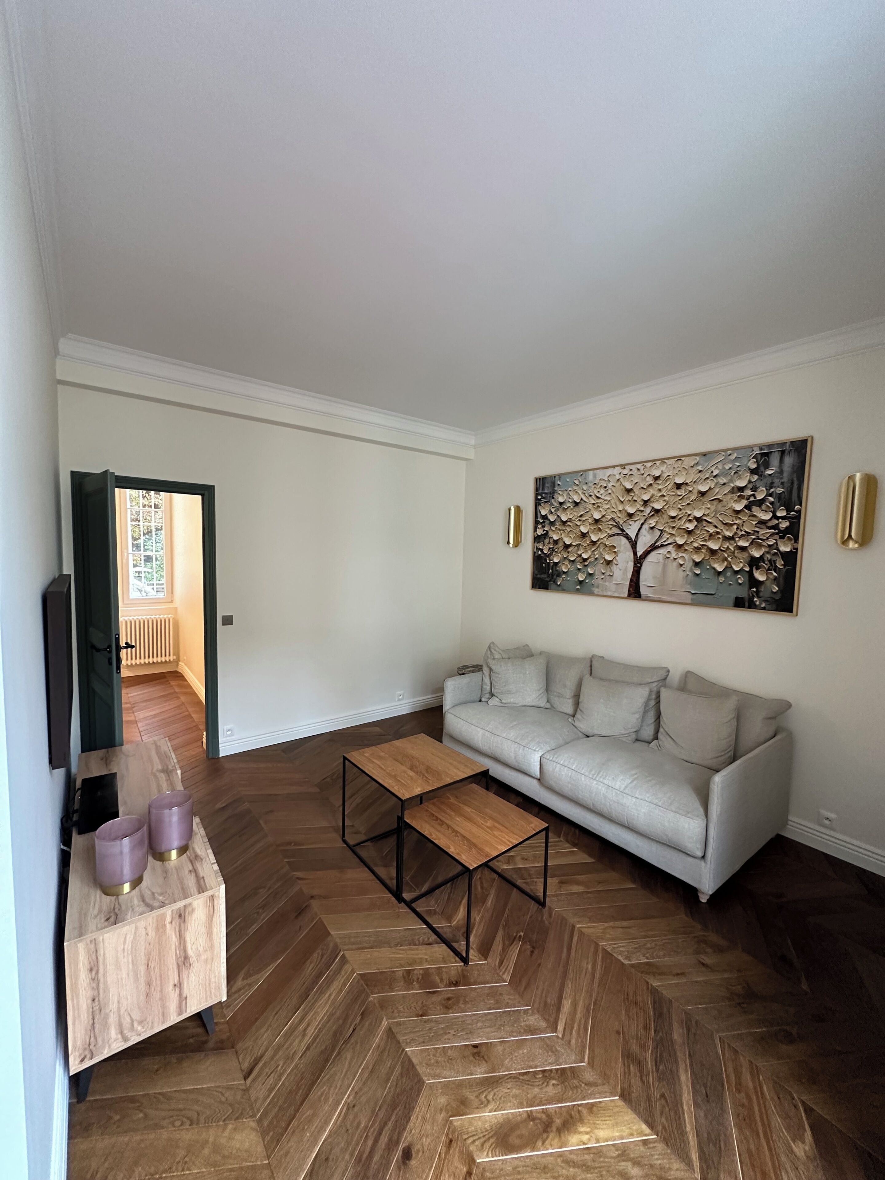 A living room with a herringbone floor, a grey sofa, a wooden coffee table, and a painting of a tree.