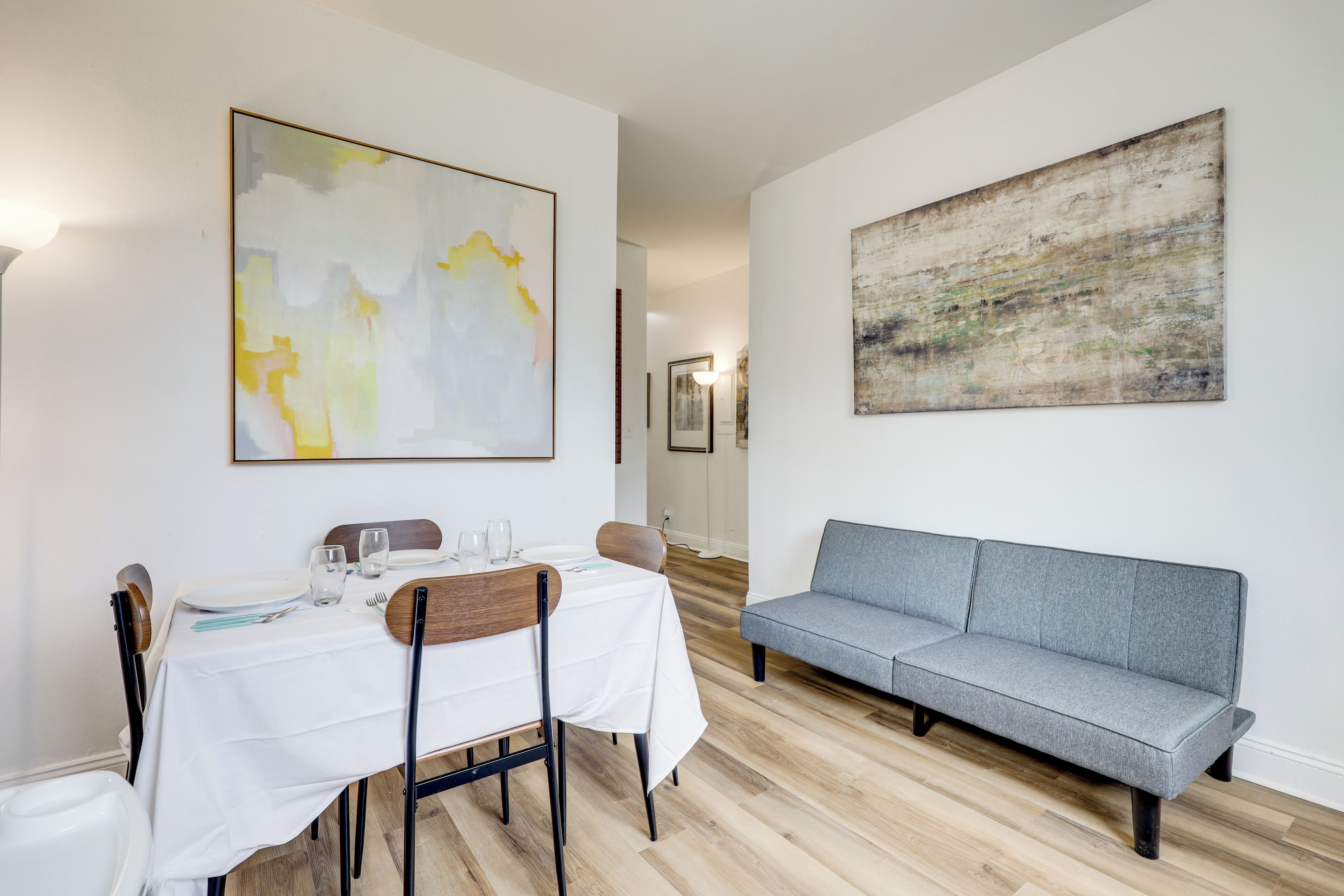 A dining area with a table set for two, a grey sofa, and abstract wall art.