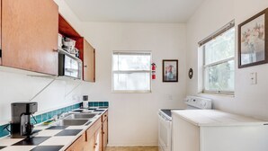 Eine kleine Küche mit Mikrowelle, Kaffeemaschine und Geschirrspüler. Ein Fenster mit Jalousien, ein Feuerlöscher und ein gerahmtes Bild sind an der Wand zu sehen.