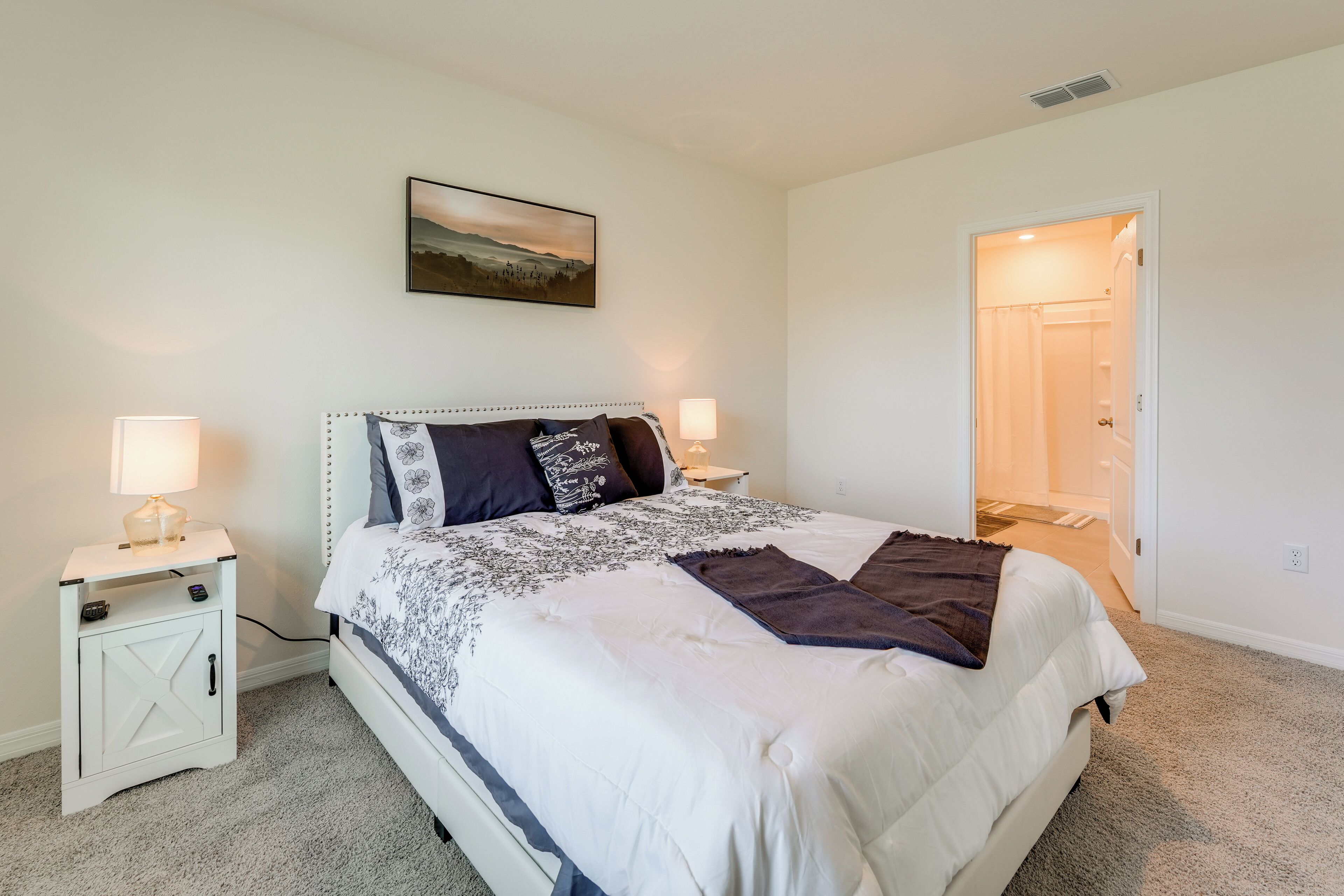 A bedroom with a bed, nightstands, a lamp, a picture on the wall, and an open door to another room.