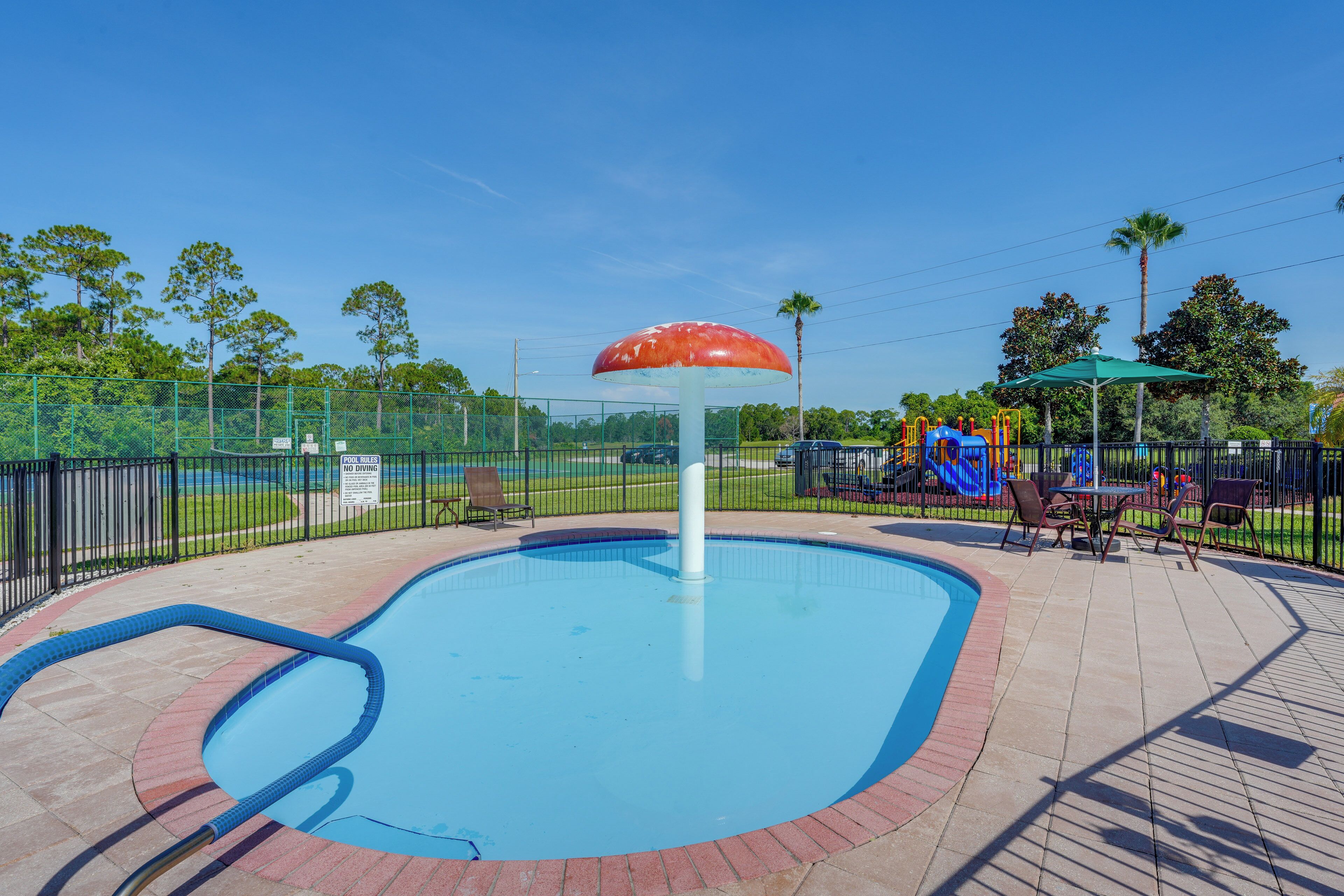 A swimming pool with a central fountain, surrounded by a fenced area with playground equipment and seating.