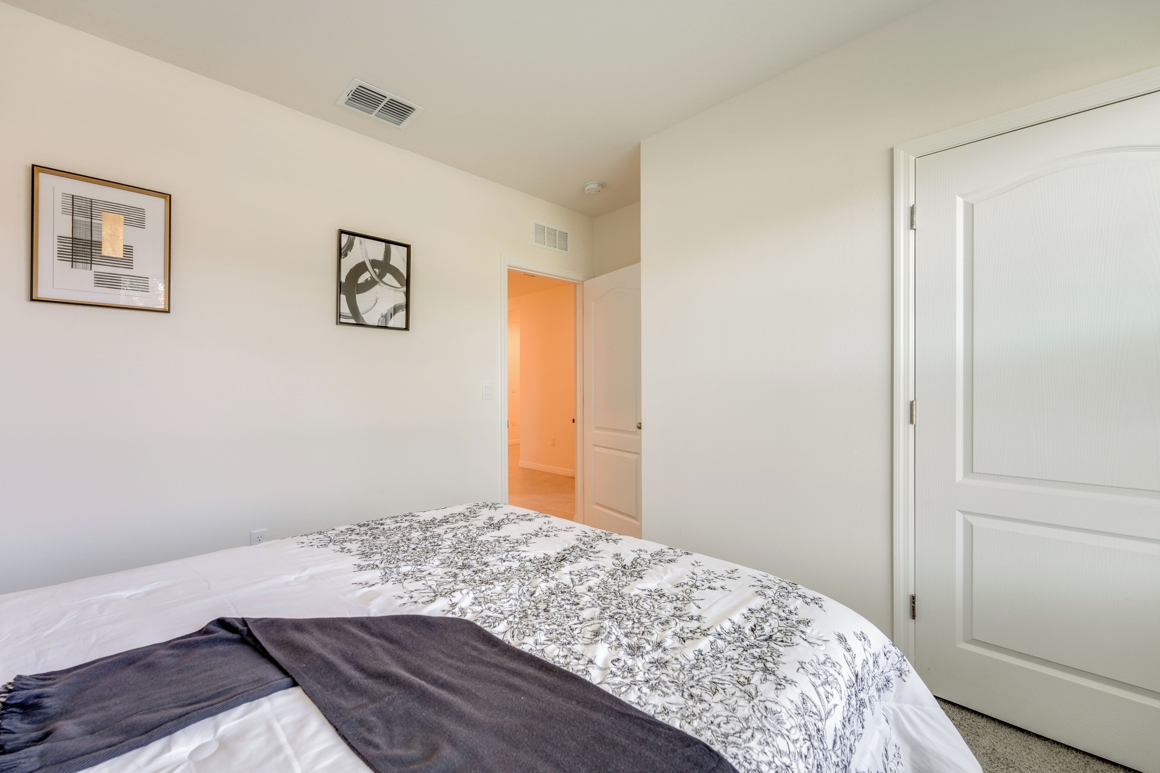 A bedroom with a bed, two framed artworks, a white door, and a hallway leading to another room.