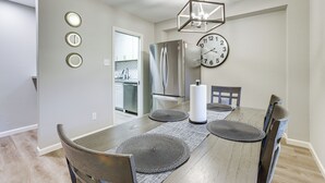 Un comedor con una mesa de madera, cuatro sillas, una araña, un reloj y un refrigerador al fondo.
