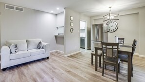 Una sala moderna con un sofá de color claro, un comedor con una mesa y sillas de madera, y una cocina al fondo.