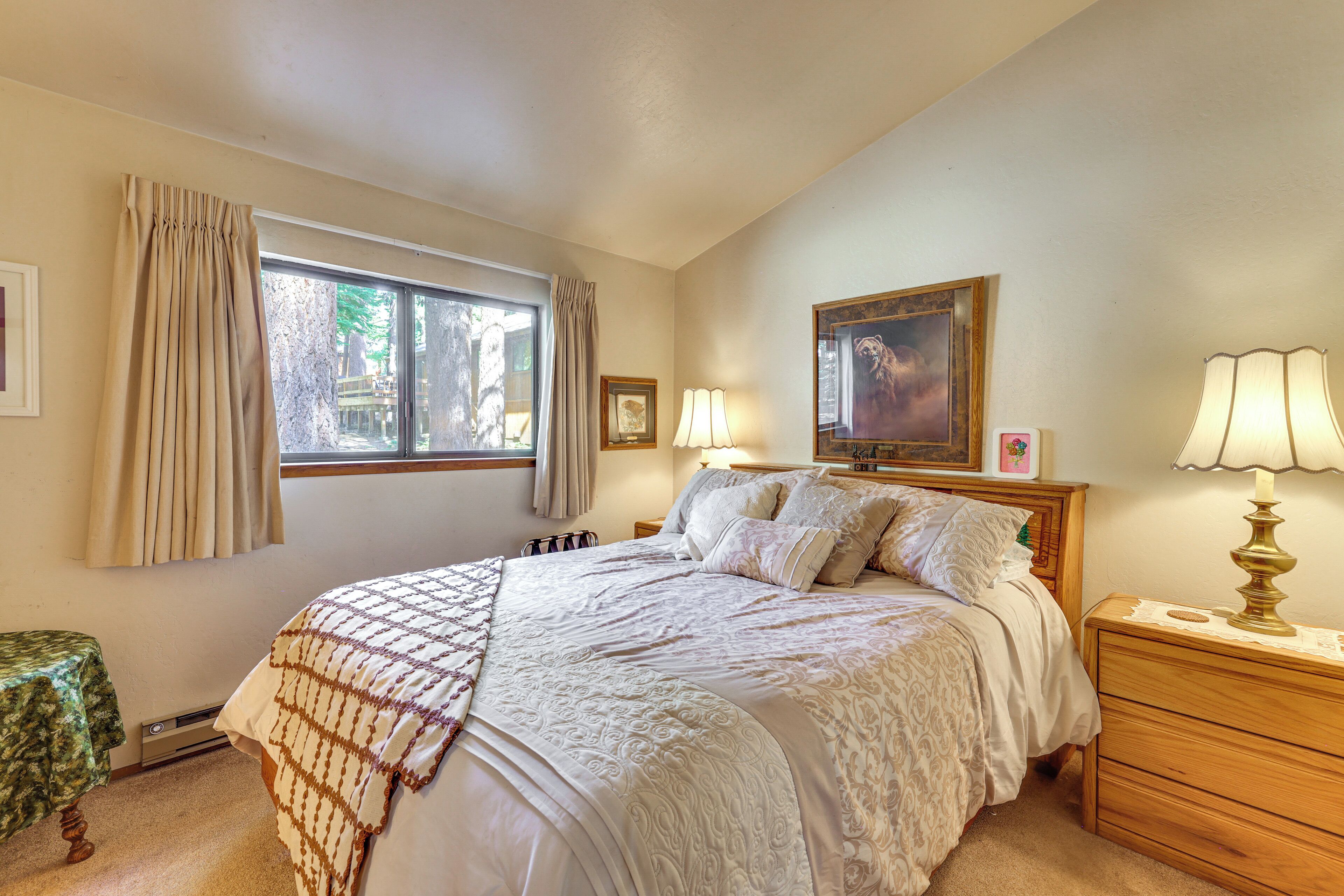 A bedroom with a large bed, a nightstand with a lamp, a window with curtains, and a framed picture on the wall.