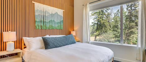 Un dormitorio con una cama grande, pared de paneles de madera, un cuadro, una mesita de noche con lámpara y una ventana con vistas a árboles.