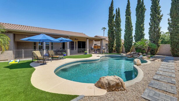 A swimming pool with a curved design, surrounded by a pebble walkway, and a patio area with chairs and umbrellas. - Extended-stay Oasis: Gilbert Getaway w/ Office! (Gilbert)