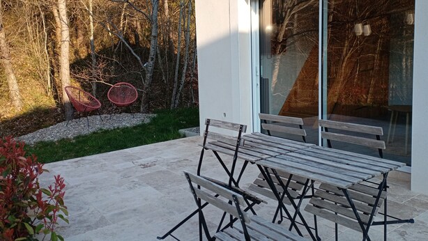 Um pátio externo com mesa e bancos de madeira, rodeado por um jardim com árvores e uma cadeira vermelha.