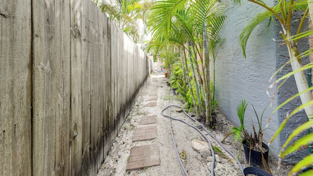 Un sendero estrecho bordeado por una valla de madera y un muro, con plantas en macetas y una manguera a lo largo del camino.