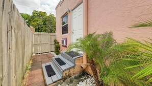 Un estrecho callejón con una cerca de madera, un edificio rosa con una puerta blanca y un pequeño jardín con plantas de palma.
