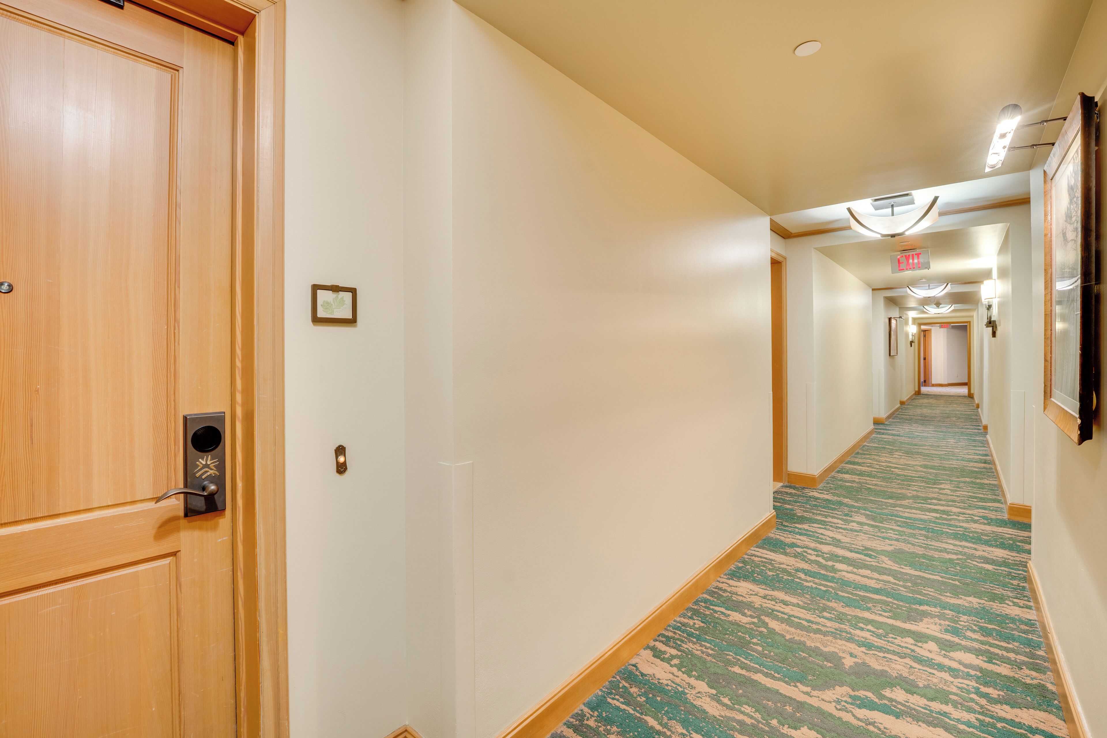 Un couloir d’hôtel avec des portes en bois, une moquette à motifs et un long couloir éclairé par un luminaire au plafond.