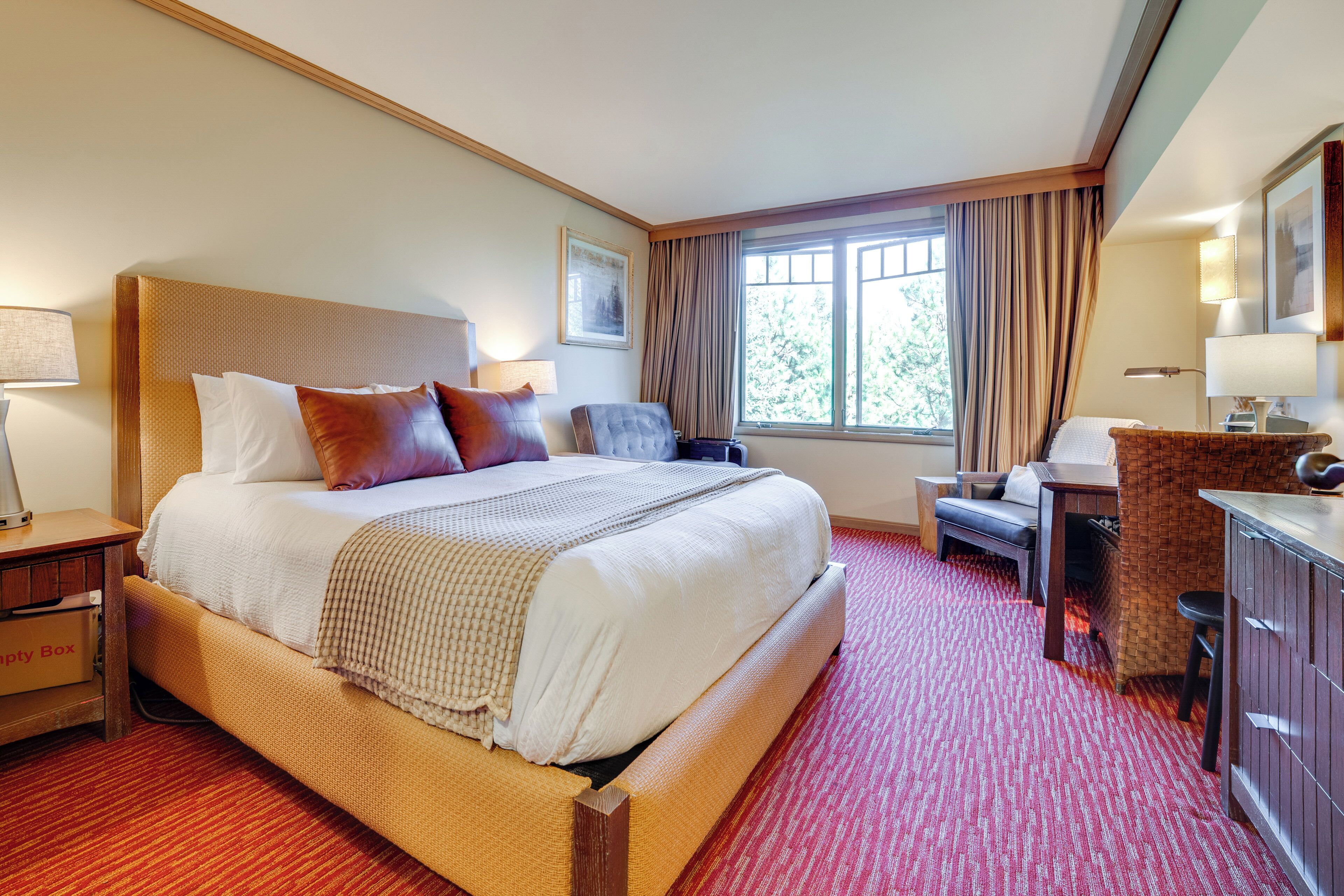 A hotel room with a large bed, a desk, a chair, a small table, and a window with curtains.