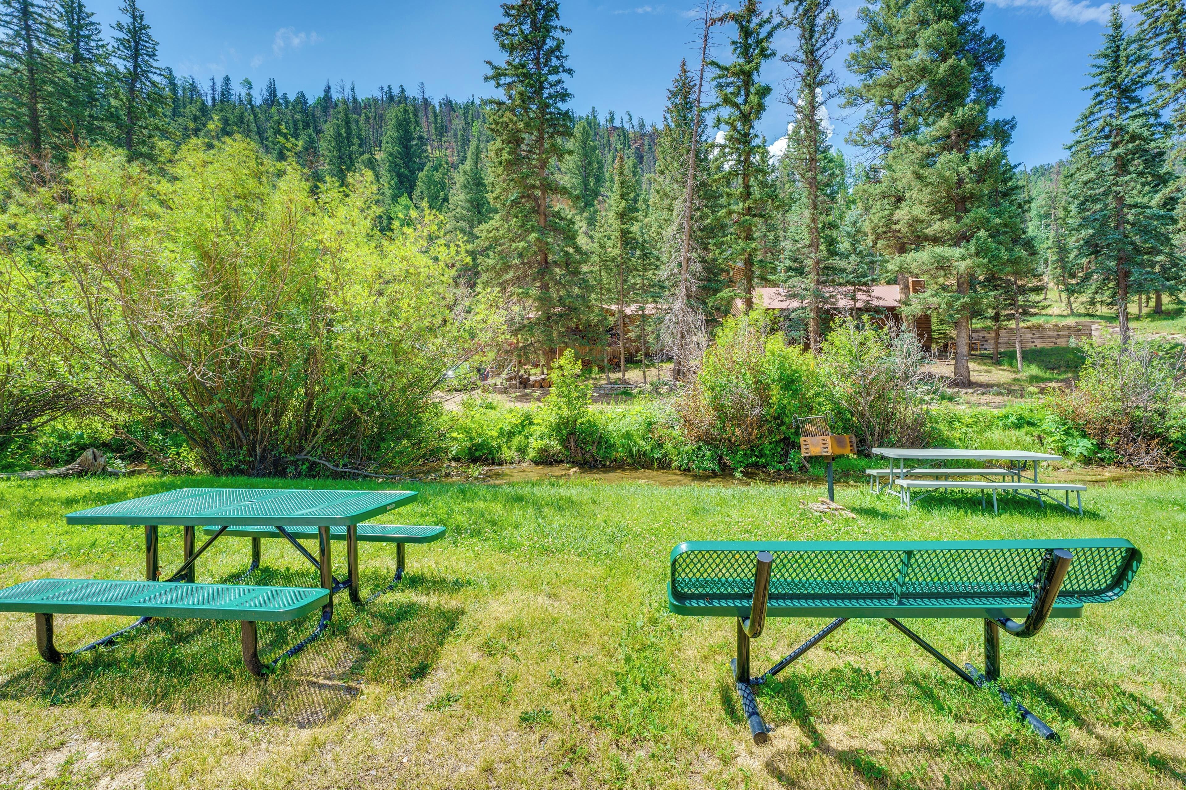 A picnic area with green tables and benches, surrounded by trees and a small building in the background.
