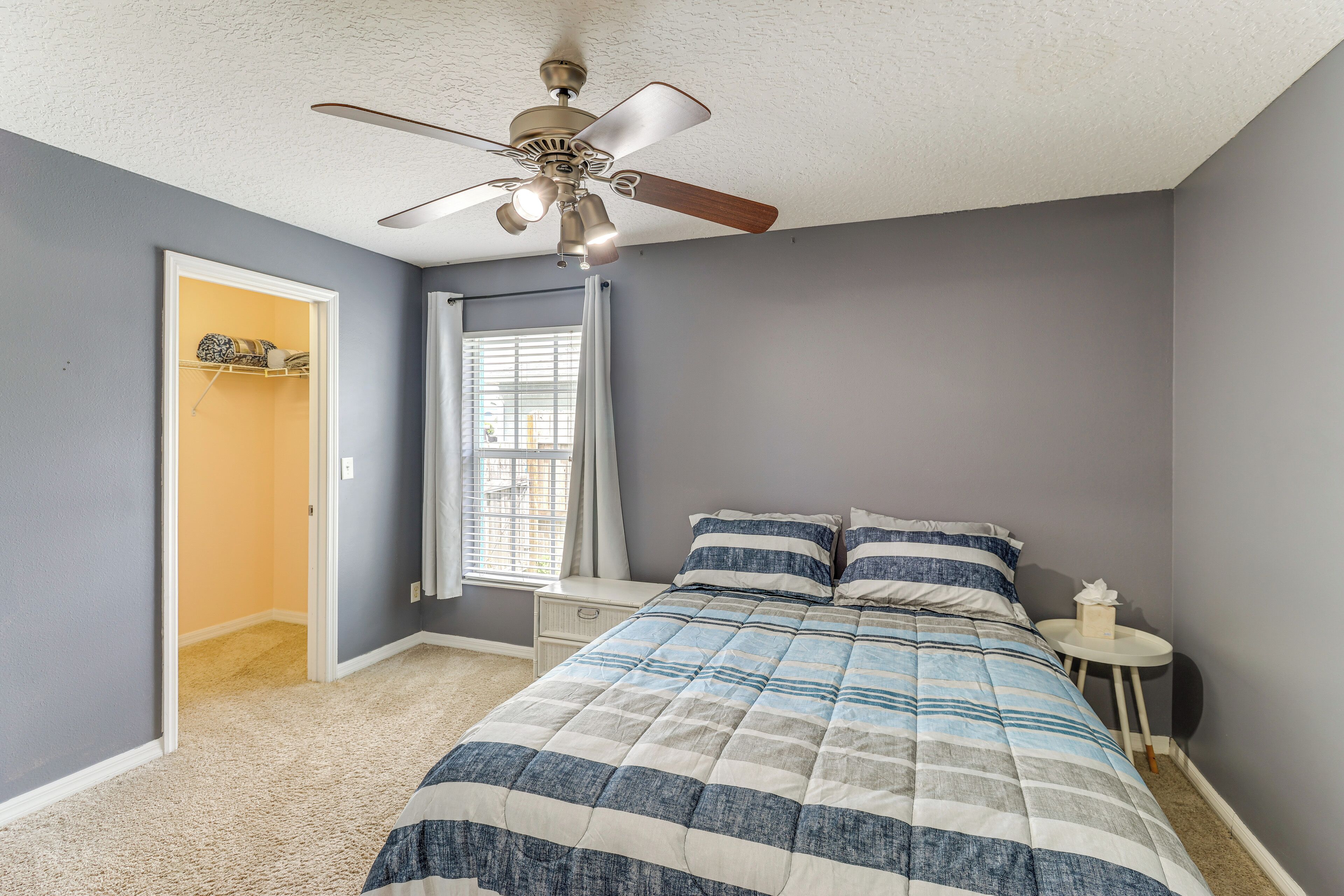 A bedroom with a bed, a nightstand, a ceiling fan, a window with curtains, and a door leading to another room.