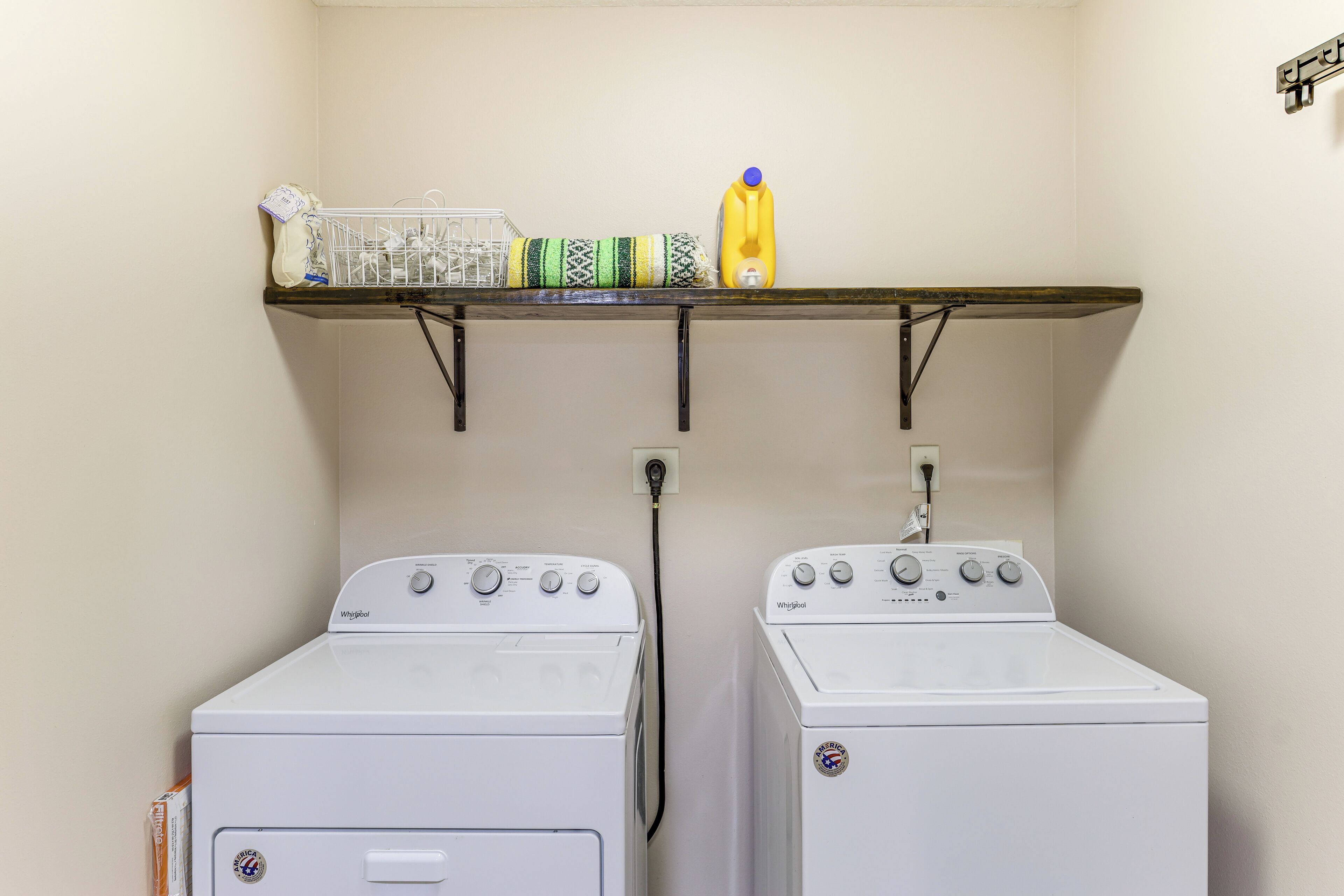 A laundry area with two white Whirlpool washers, a shelf above holding a basket, detergent, and a spray bottle.