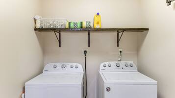 A laundry area with two white Whirlpool washers, a shelf above holding a basket, detergent, and a spray bottle.