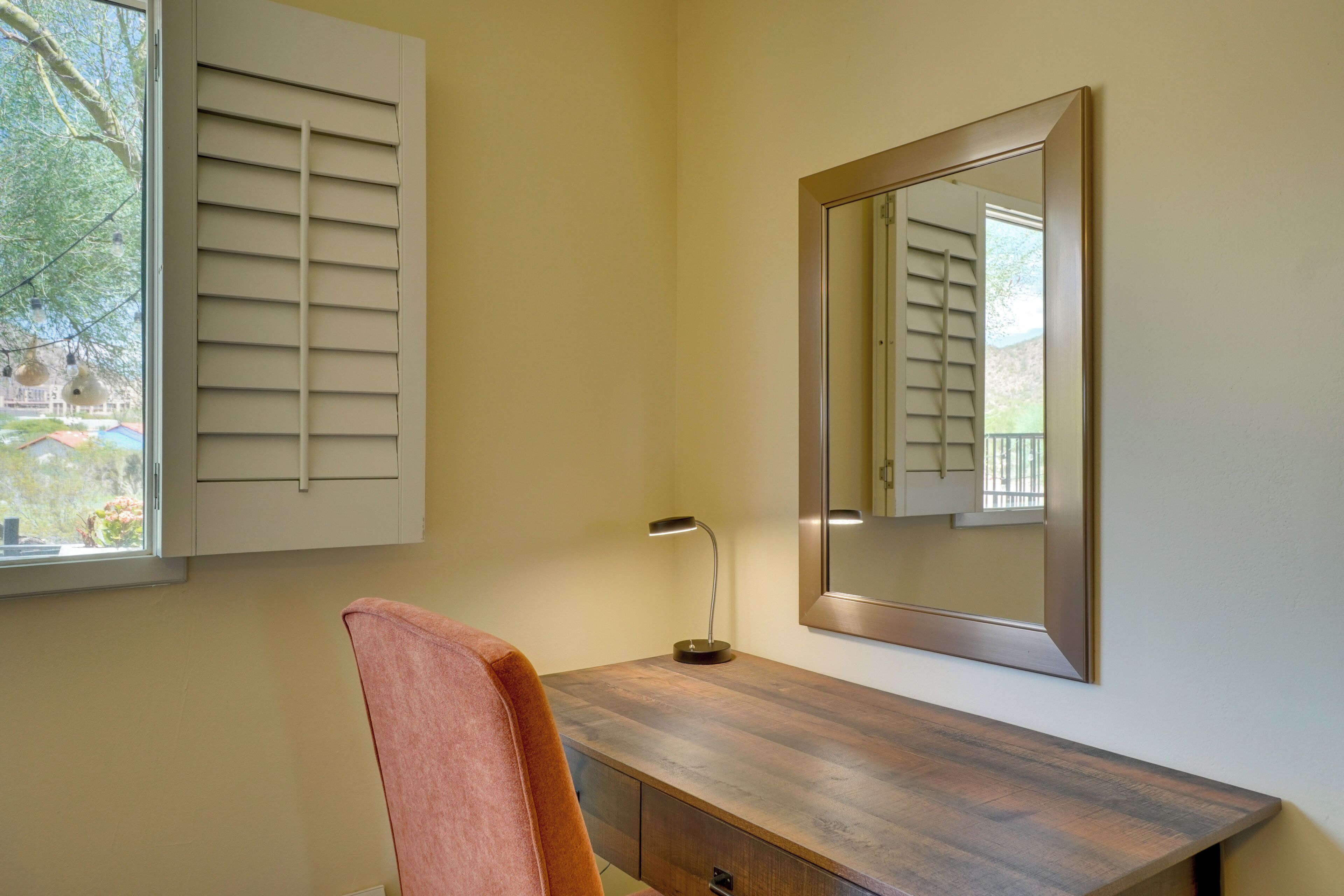 Une pièce avec un bureau en bois, une chaise, une lampe, un grand miroir et une fenêtre avec des volets.