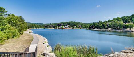 A serene lakeside view with a walking path, rocky shoreline, and buildings in the background.