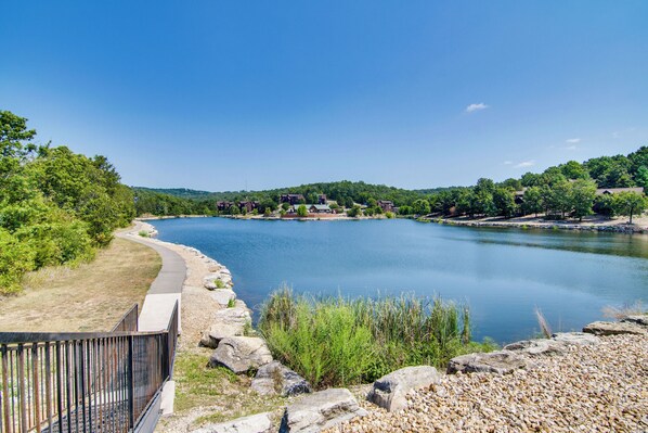 A serene lakeside view with a walking path, rocky shoreline, and buildings in the background. - Charming Golf Resort Oasis Near Silver Dollar City (Branson West)