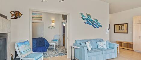Ein Wohnzimmer mit einem blauen Sofa, einem weißen Korbstuhl und einem kleinen Tisch. An den Wänden befinden sich Wandbilder, darunter ein Fisch und ein Seepferdchen. Eine Tür führt in einen anderen Raum.