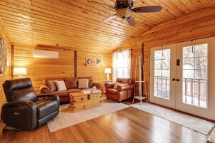 Interior de una cabaña de troncos con techo de madera, un sillón de cuero, un sofá, un cofre de madera y un ventanal corredizo que da al exterior.