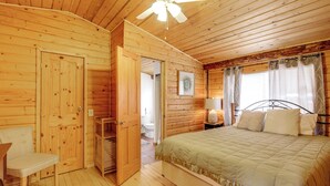 A wooden cabin bedroom with a bed, a chair, a nightstand with a lamp, and a ceiling fan.