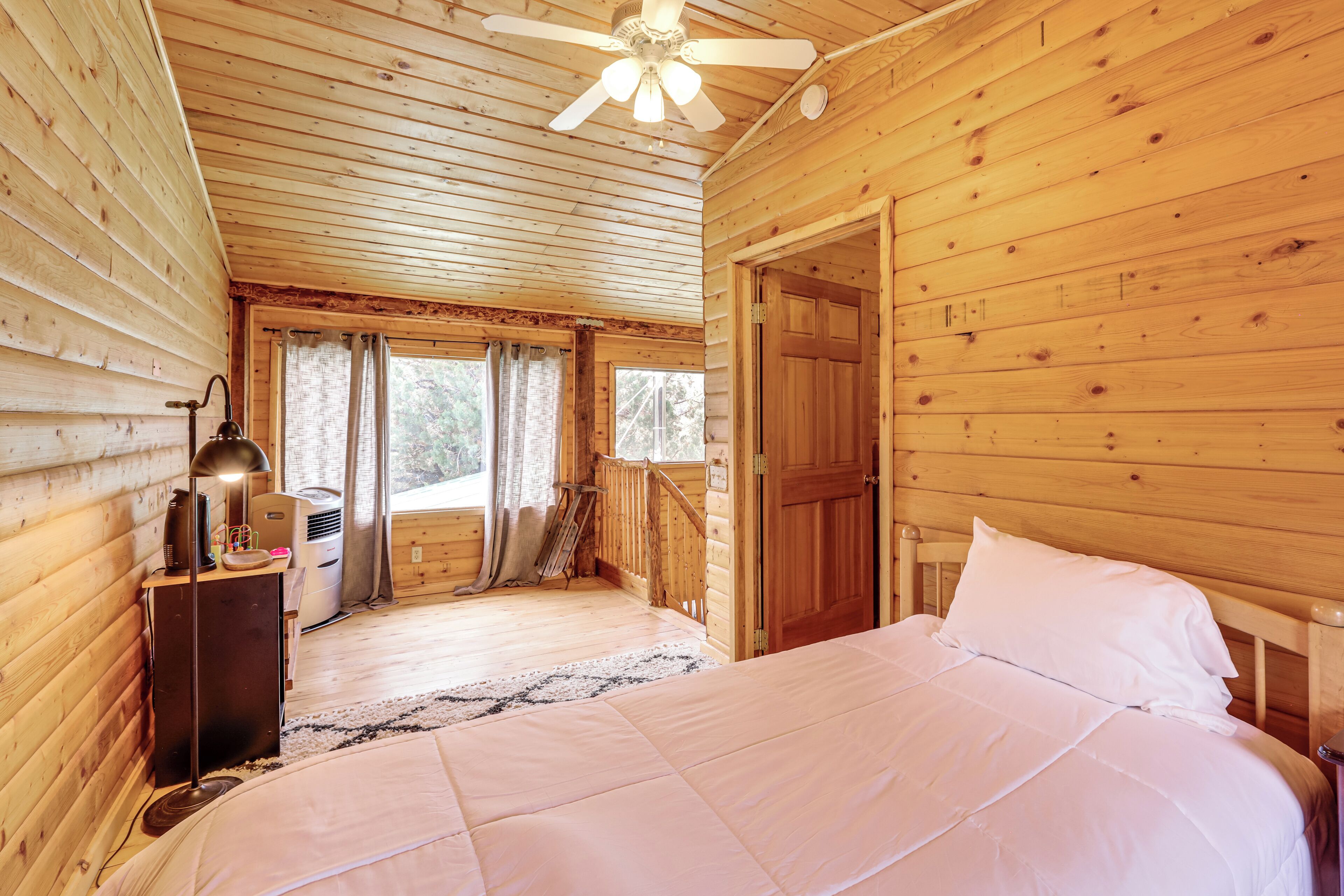 A wooden cabin bedroom with a bed, a ceiling fan, a nightstand, and a window with curtains.