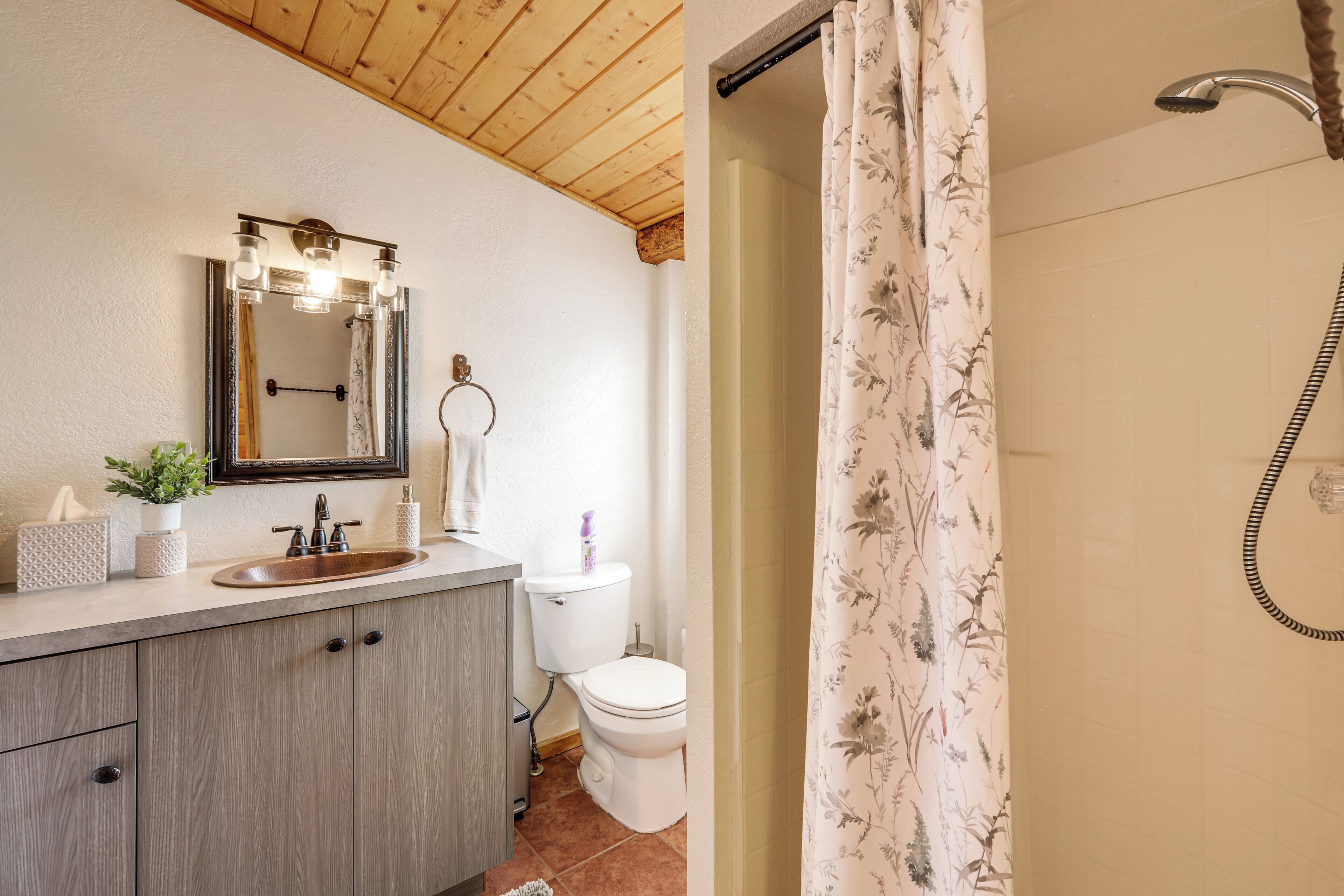 A bathroom with a wooden ceiling, a sink with a mirror, a toilet, and a shower with floral curtains.