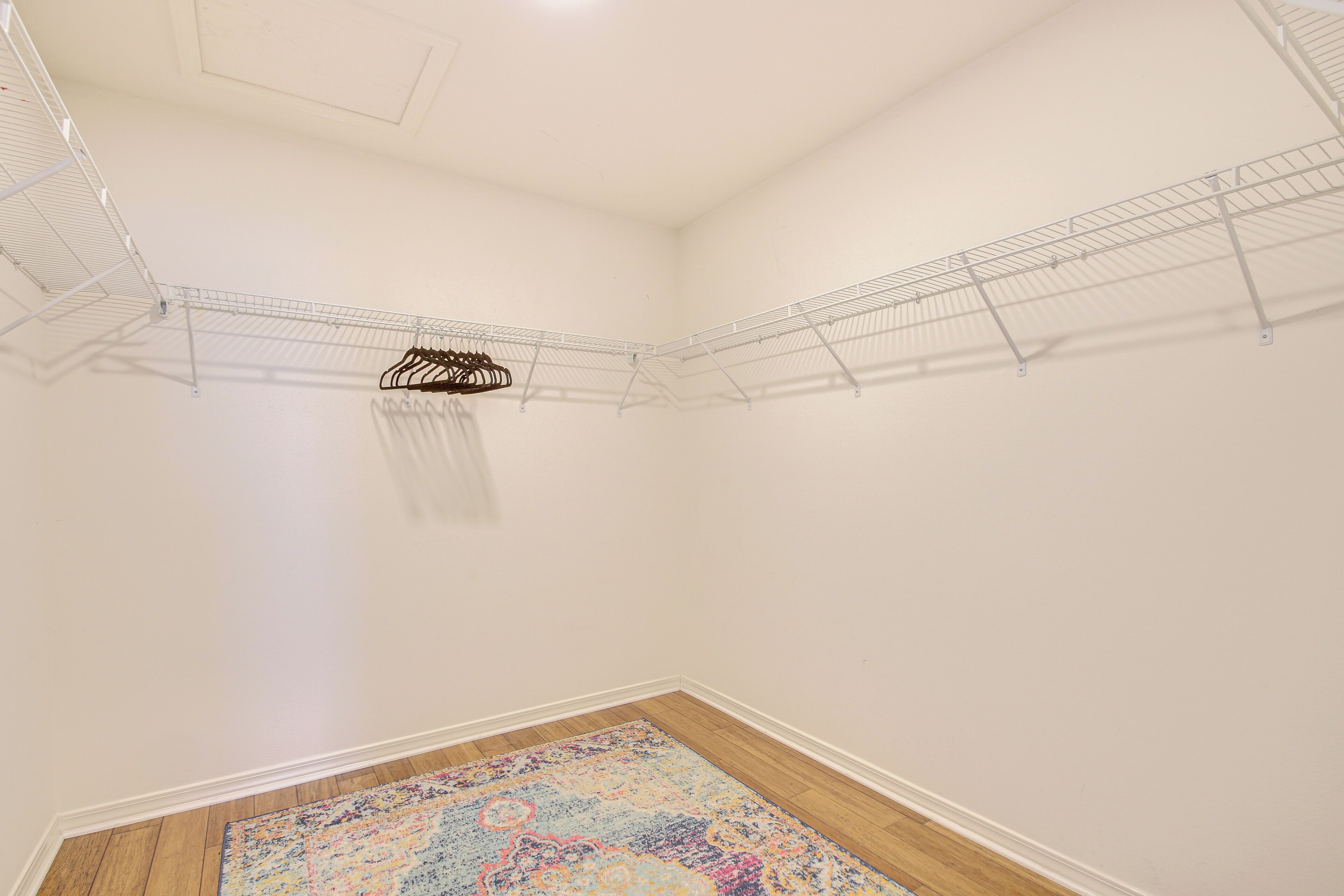 Una habitación con piso de madera, paredes blancas y un tapete colorido. Se observan dos estanterías metálicas en las paredes.