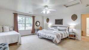Ein Schlafzimmer mit einem großen Bett, einem kleineren Bett, einem Schreibtisch und einem Stuhl. Ein Fenster mit Vorhängen, ein Deckenventilator und ein wandmontierter Spiegel sind ebenfalls vorhanden.