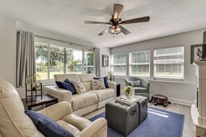 A spacious living room with a beige leather sofa, a gray ottoman, and a blue rug. There are large windows with blinds, a ceiling fan, and a fireplace. - Withlacoochee River Retreat w/ Dock: Paddle & Fish (Citrus Springs)